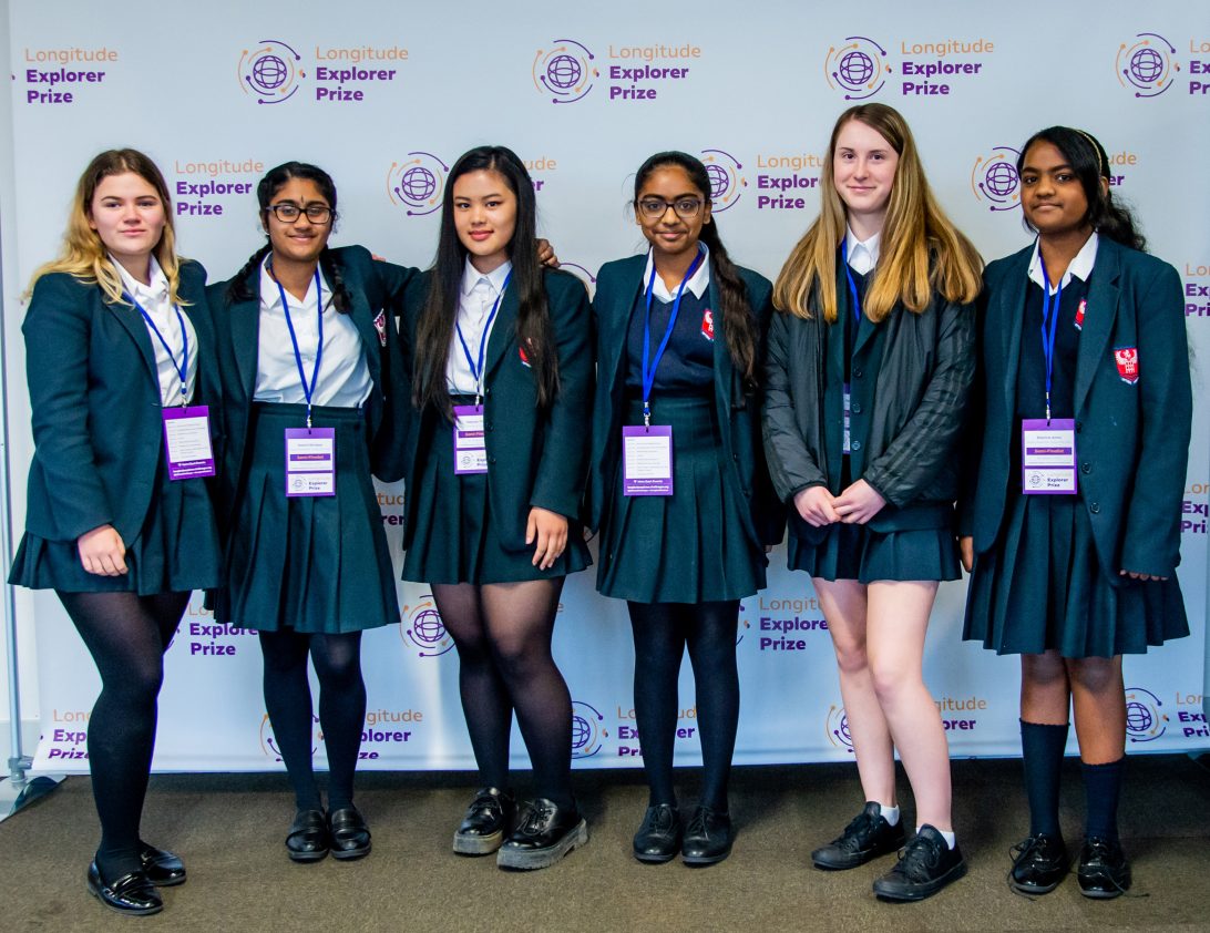 Six teenagers who present as women in a navy blue school uniform, standing in front of a roll up banner featuring the 2020 Longitude Explorer Prize logo
